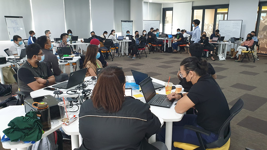 Group in a workshop with laptops, masked, listening to a presenter in a modern office.