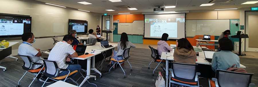 Participants in a hybrid workshop watching a presentation on a large screen and monitors.