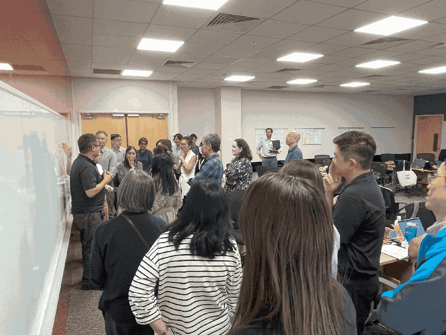 A group of workshop participants gathered around a whiteboard, listening to a presentation.